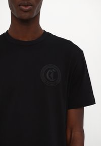 Camiseta de algodón negra con un logo redondo en un tono más oscuro, ubicado en el área del pecho izquierdo, con un clásico escote redondo.