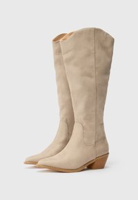 Bottes en daim beige jusqu'au genou avec un bout pointu, une texture douce et un talon en bois empilé, présentant un design simple et épuré sans ornements.