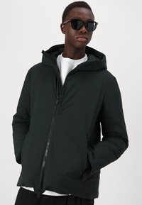 Dark green puffer jacket with a hood, front zipper closure, and side pockets. Smooth texture and contemporary design. Paired with sunglasses.