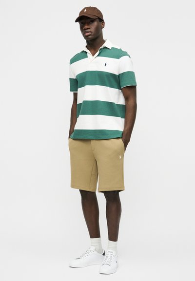 Άνδρας που στέκεται, φορά καφέ καπέλο, κοντομάνικο πόλο με πράσινες και λευκές ρίγες, μπεζ σορτς, λευκές κάλτσες και λευκά αθλητικά παπούτσια.