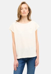 Helles beige T-Shirt mit kurzen Ärmeln und rundem Ausschnitt, weicher Textur und dezentem Logo-Detail auf der linken Brust. Kombiniert mit blauen Jeans.