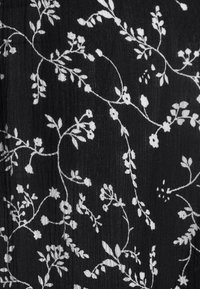 Tissu noir avec broderie florale blanche représentant des vignes et des fleurs, affichant des motifs complexes et une surface texturée.
