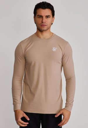 Mand med mørkt hår iført en beige langærmet skjorte med et hvidt logo på venstre bryst, stående mod en ensartet lys baggrund.