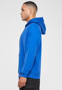 Sudadera de algodón azul con bolsillo canguro, capucha con cordón y puños acanalados. La vista lateral muestra un ajuste relajado y una textura suave.