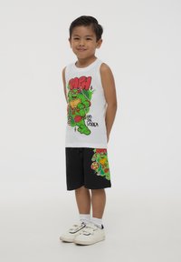 Maglietta senza maniche bianca con una grafica di un supereroe verde, abbinata a pantaloni corti neri con una grafica simile. Scarpe bianche con strappi in velcro.