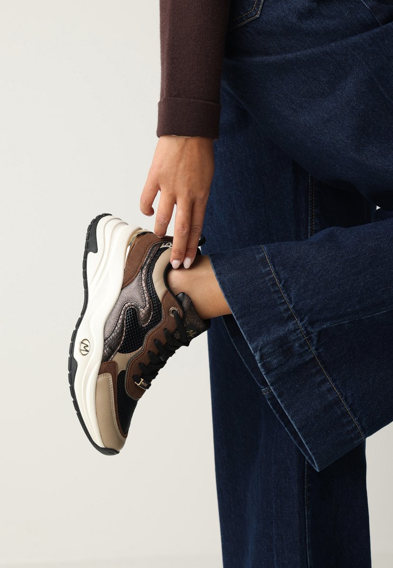 Tenisky z hnědého síťovaného materiálu a kůže s texturovanými akcentovými panely. Mají černé šněrování a masivní bílou podrážku. Přítomný detail loga na boku.