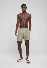 Mønstrede badetights i olivengrøn med elastisk talje og sidelommer. Kombinert med lyse sandaler. Glatt stofftekstur.