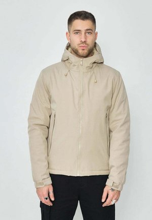 Homme portant une veste à capuche beige avec une fermeture éclair et des cordons, debout devant un fond uni clair.