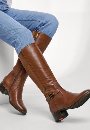 Personne portant des bottes en cuir marron hauteur genou avec fermetures éclair latérales et détail de boucle, associées à un jean bleu, assise sur une surface blanche.