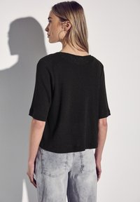 Maglietta nera a maniche corte in maglia con una vestibilità comoda. Presenta un ampio scollo e un tessuto testurizzato. Indossata con jeans grigi chiari e usurati.