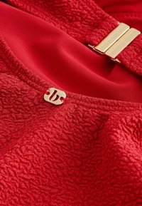 Tessuto rosso testurizzato con una piccola targhetta logo dorata e una fibbia rettangolare dorata attaccata a una fodera in rete rossa liscia.
