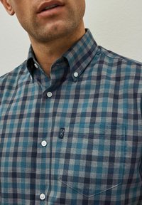 Camisa de botones con un patrón a cuadros en azul y gris, con un bolsillo en el pecho y detalle del logo. Hecha de una tela con textura suave.