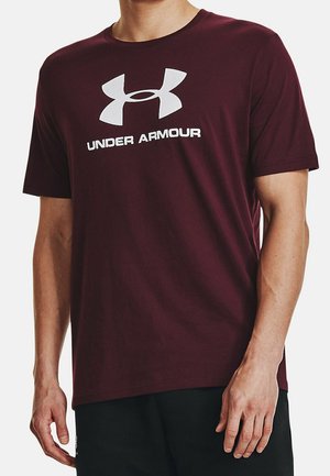 Mann trägt ein bordeauxfarbenes Under Armour T-Shirt mit großem weißem Logo und Text auf der Brust, kombiniert mit schwarzen Hosen.