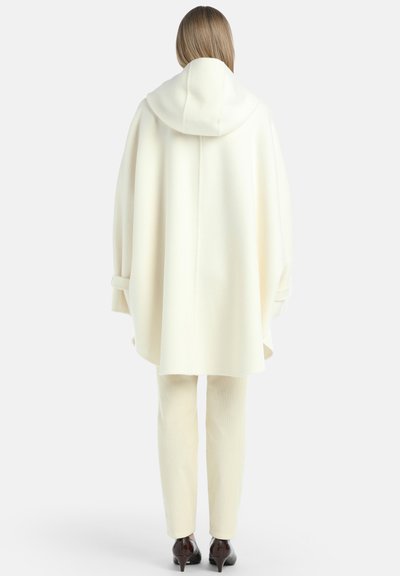 Crème-kleurige wollen poncho met een capuchon, voorzien van een ontspannen pasvorm en zijsplitten, gecombineerd met bijpassende crème broeken en hoge schoenen.