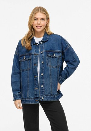 Jonge vrouw met blond haar, gekleed in een oversized blauwe spijkerjas, wit shirt en zwarte broek, staand met één hand in de zak.