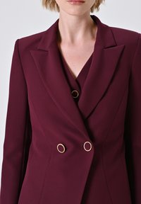Femme portant un blazer ajusté bordeaux avec de larges revers et des boutons ronds dorés, vue de la moitié de la poitrine au cou.