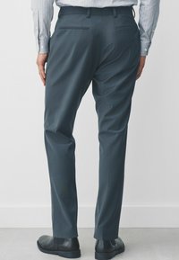 Graue maßgeschneiderte Hosen mit glatter Textur, ausgestattet mit zwei Gesäßtaschen, getragen mit schwarzen Schuhen, mit einem geraden Bein-Design und subtilen Falten.