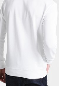 Chemise blanche à manches longues avec poignets et ourlet côtelés, présentant une texture lisse et un design simple. La vue de dos ne montre aucun graphisme ni embellissement.