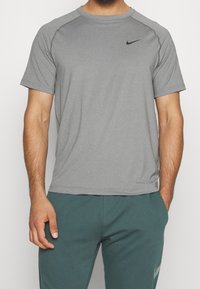 Hombre vistiendo una camiseta deportiva corta de Nike gris y pantalones de Nike en color teal, con una mano en el bolsillo y la otra relajada a un lado.