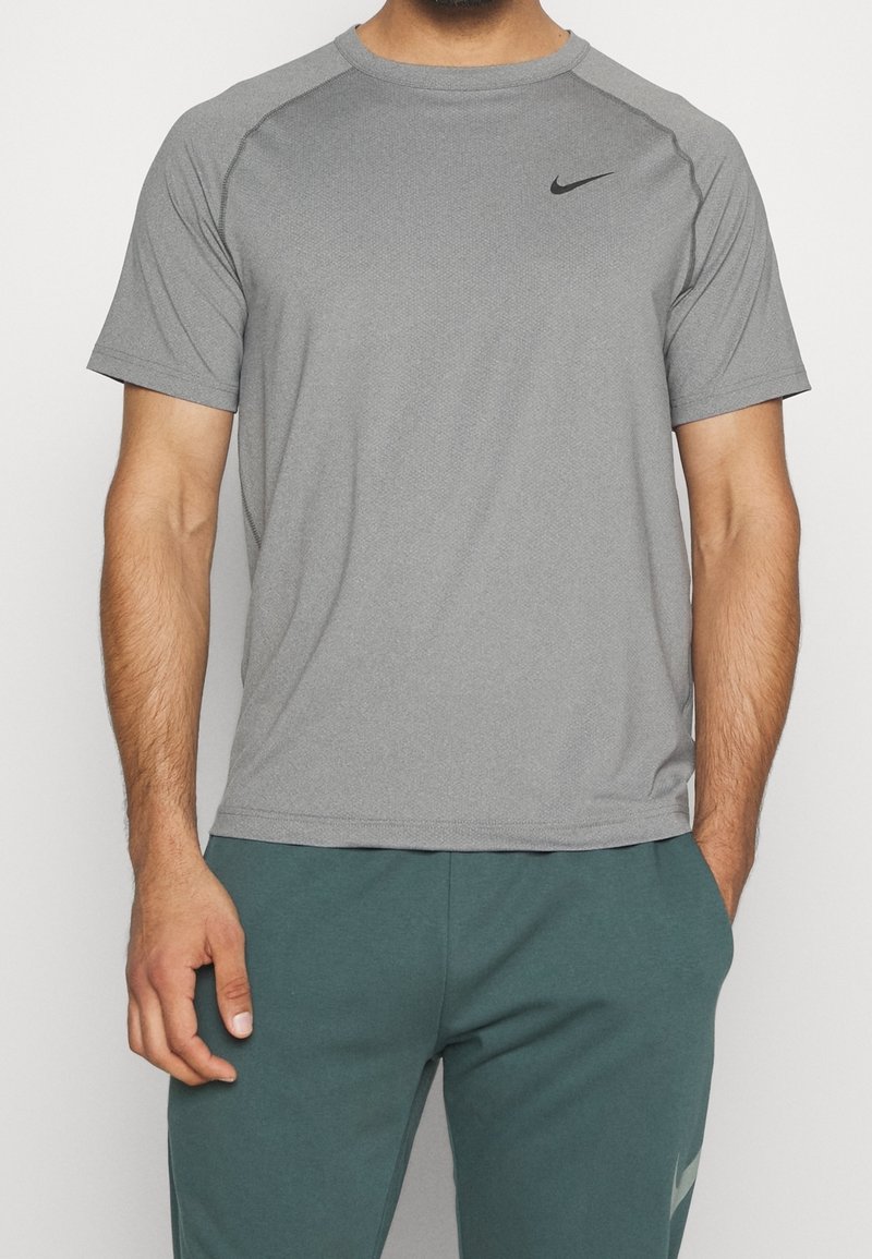Hombre vistiendo una camiseta deportiva corta de Nike gris y pantalones de Nike en color teal, con una mano en el bolsillo y la otra relajada a un lado.