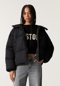 Giacca nera imbottita con zip e cappuccio, caratterizzata da un design trapuntato e polsini elasticizzati, abbinata a un maglione nero e jeans grigi.