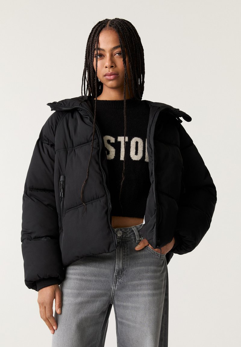 Giacca nera imbottita con zip e cappuccio, caratterizzata da un design trapuntato e polsini elasticizzati, abbinata a un maglione nero e jeans grigi.