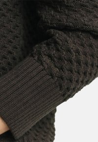 Dunkelbraune Strickpulloverärmel zeigen ein strukturiertes Design und einen gerippten Abschluss, der verschiedene Stichmuster zur visuellen Auflockerung präsentiert.