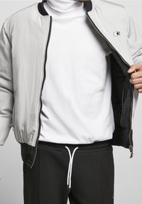 Man draagt lichtgrijs jack met rits over een witte coltrui en zwarte broek met wit trekkoord tegen een effen achtergrond.