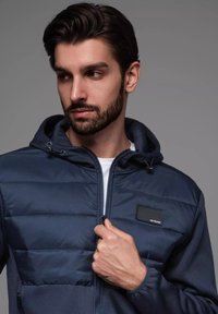 Marinejacka med vadderad front, texturerade ärmar, kapuschong och sidfickor. Har en liten logotyp på bröstområdet. Dragkedjestängning.