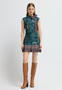 Kurzes Kleid in Türkis mit Paisleymuster und einer Schleifenkragen-Applikation. Strukturiertes Material, ausgestellter Saum, kombiniert mit hohen braunen Stiefeln mit Absatz.