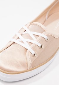 Chaussure décontractée en tissu beige avec lacets blancs entrecroisés, œillets métalliques et semelle en caoutchouc blanche avec bordure beige.
