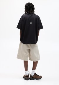 Schwarzes Übergröße-T-Shirt mit dem Text "Eine neue Idee" auf dem Rücken, kombiniert mit beigen Shorts und braunen Sportschuhen. Weiße Kniesocken sind sichtbar.