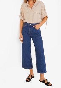 Camisa beige de manga corta y cuello, con dos bolsillos en el pecho, combinada con vaqueros de mezclilla azul de pierna ancha y cintura alta, y sandalias negras.