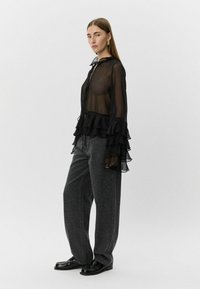 Black sheer blouse with frilled sleeves; paired with gray leopard print pants. Model wears black shoes. Minimalist background.