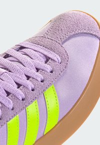 Fioletowy but zamszowy i tekstylny wierzch, z trzema neonowozielonymi paskami, teksturowanymi sznurowadłami i brązową gumową podeszwą.