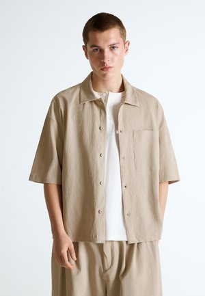 Ung mand iført beige kortærmet tekstureret skjorte med knapper over en hvid t-shirt og matchende beige bukser, stående mod en hvid baggrund.