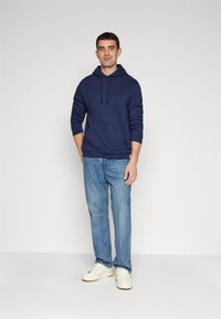Hanorac cu glugă de culoare albastru marin, dotat cu buzunar frontal, asortat cu blugi de culoare albastră deschisă și adidași albi. Design simplu, material din bumbac, croială lejeră.
