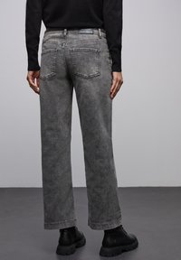 Jeans gris à jambe large avec une finition délavée, dotés de deux poches arrière et de coutures visibles. Portés avec des bottines noires.