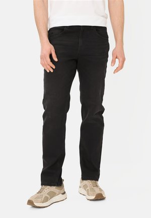 Mann trägt schwarze Jeans und beige Turnschuhe, steht vor einem schlichten weißen Hintergrund.