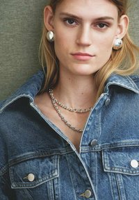 Giacca di denim con bottoni argentati, collana a catena argentata stratificata e grandi orecchini circolari argentati. Lo sfondo neutro esalta le caratteristiche.