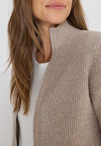 Beige rippad kardi med ståkrage, med dragkedja och mjuk textur. Bäres över en vit skjorta, vilket visar en mysig design.