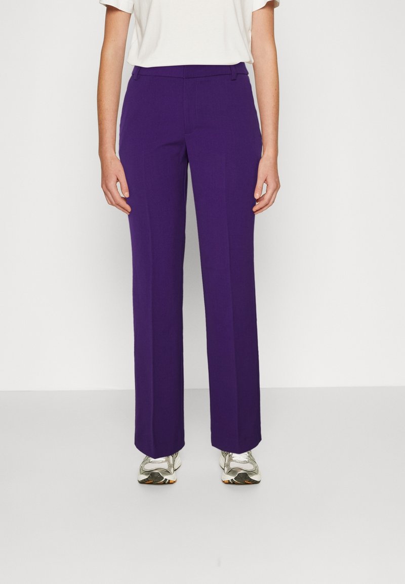 Personne portant un pantalon violet à jambes droites, une chemise blanche à manches courtes et des baskets blanches, debout sur un fond clair uni.