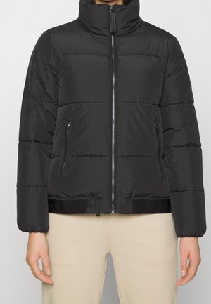 Personne portant une doudoune matelassée noire avec un col montant, une fermeture éclair à l'avant et des poches latérales zippées, associée à un pantalon beige.