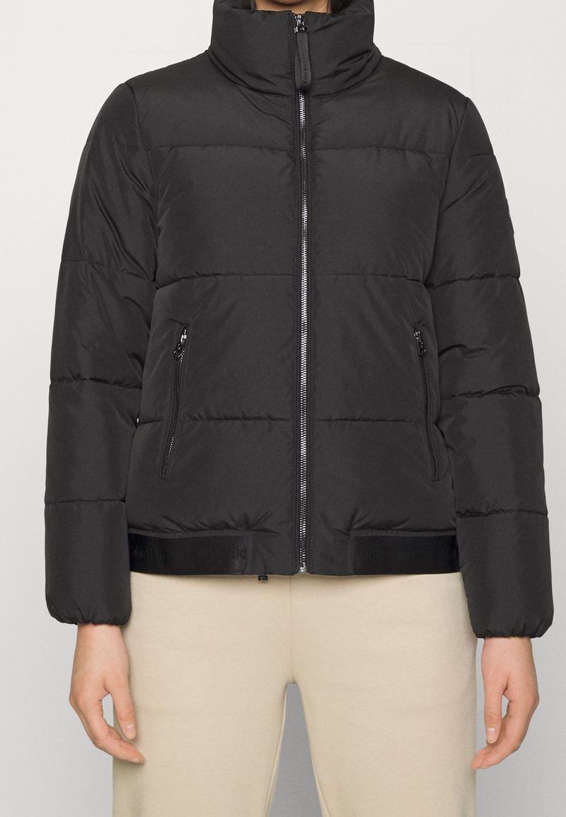 Personne portant une doudoune matelassée noire avec un col montant, une fermeture éclair à l'avant et des poches latérales zippées, associée à un pantalon beige.