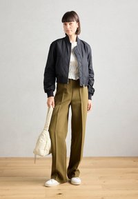 Giacca bomber nera con polsini a coste, blusa bianca testurizzata e pantaloni larghi color oliva. Tiene una borsa beige, indossa sneakers bianche su un pavimento di legno.
