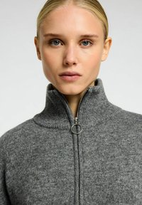 Sivi zip-up dukserica s visokim ovratnikom, teksturirano pletivo i metalna okrugla kopča za zatvaranje. Prikaz samo blizu odjevnog predmeta.