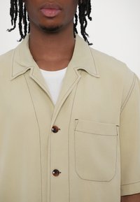 Chemise courte beige clair à col pointu, avec des boutons bruns, une poche poitrine et des détails de couture contrastés. Texture de tissu lisse.