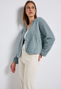 Giacca in fleece di colore blu chiaro con collo rotondo, maniche lunghe e tasche laterali. Superficie testurizzata con dettagli di rifinitura a contrasto.