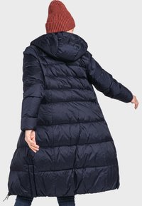 Marineblaue Steppjacke mit Kapuze, ausgestattet mit einem gesteppten Design, seitlichen Reißverschlusstaschen und einem lässigen Schnitt. Getragen über einer Jeans und einer orangefarbenen Strickmütze.