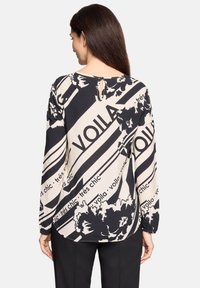 Langærmet skjorte i beige og sort med et grafisk mønster. Har en nøglehuldetalje bagpå og elastiske manchetter. Glat stoftekstur.